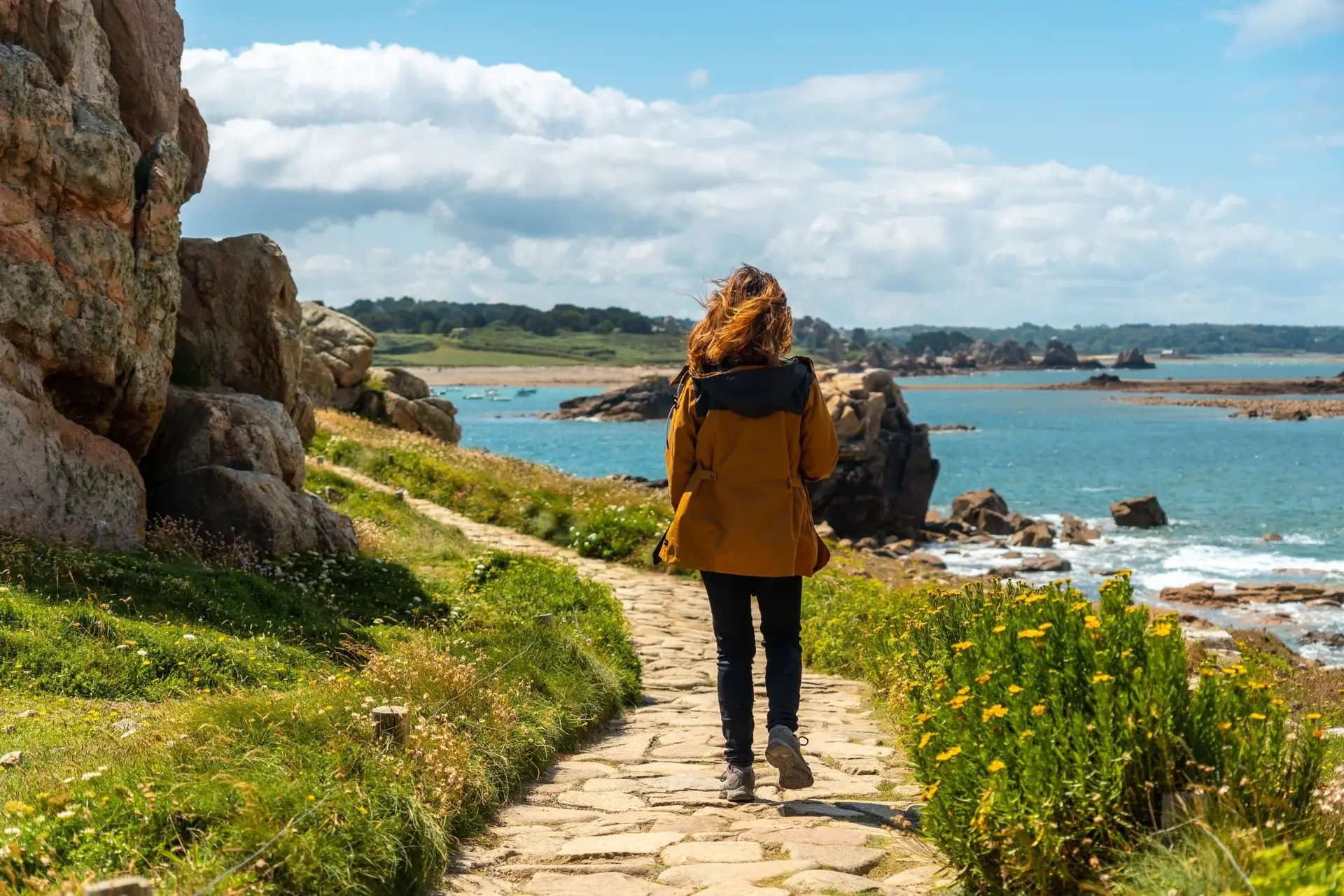 Randonneuse de dos marchant sur un sentier pavé le long d'une côte rocheuse sauvage en Bretagne, portant une veste ocre face à une mer bleu turquoise parsemée d'îlots sous un ciel ensoleillé.