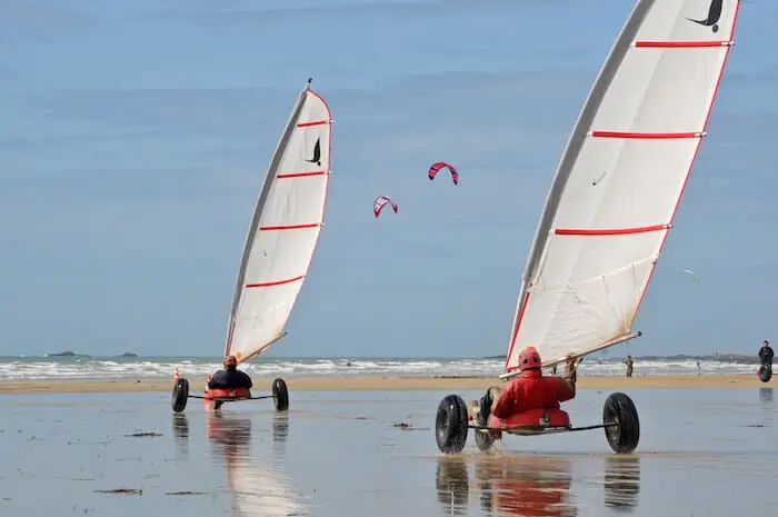 Deux chars à voile roulant à vive allure sur une plage de sable mouillé à marée basse, avec des kitesurfs en arrière-plan.