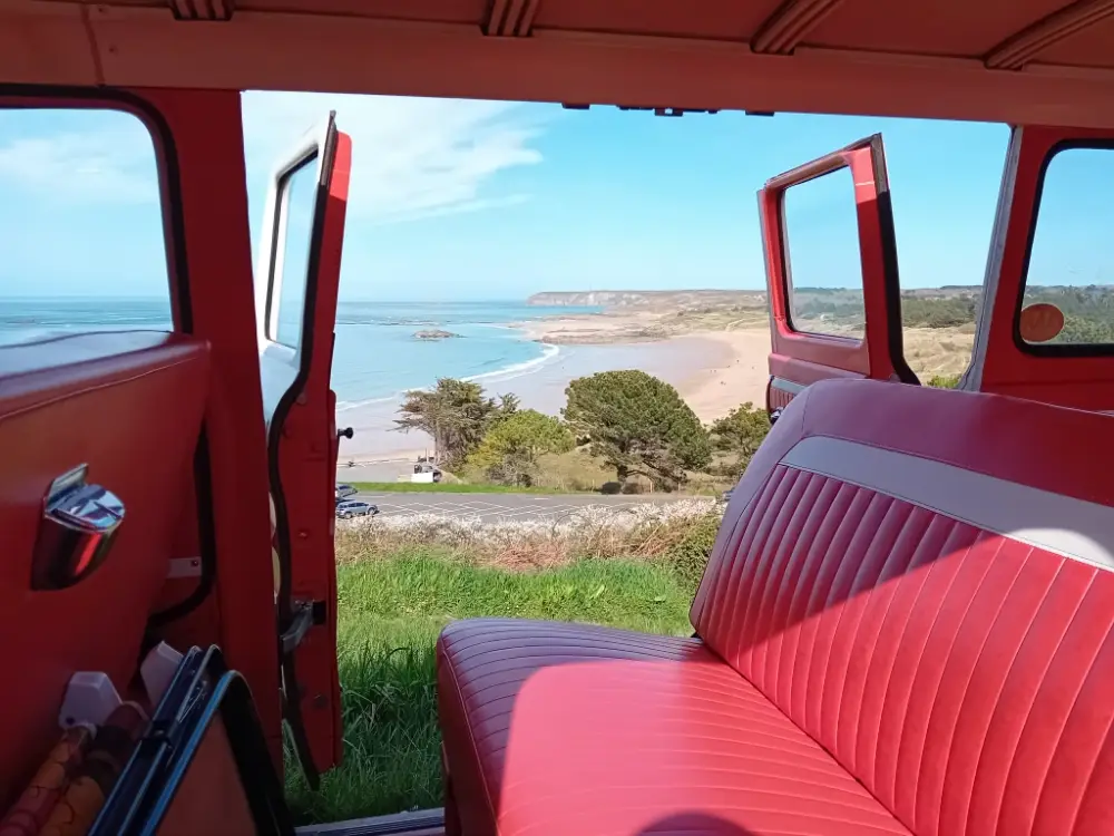 Vue depuis l'intérieur d'un van vintage aux sièges en cuir rouge, avec les portes ouvertes sur une plage et la mer.